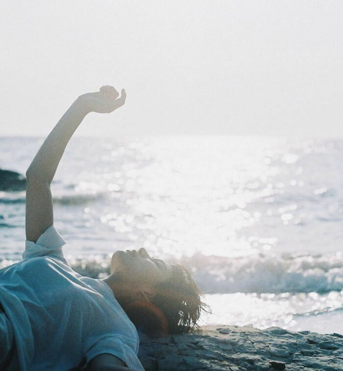 A person lying back on the sand at the water's edge, one arm raised toward a bright, hazy sea.