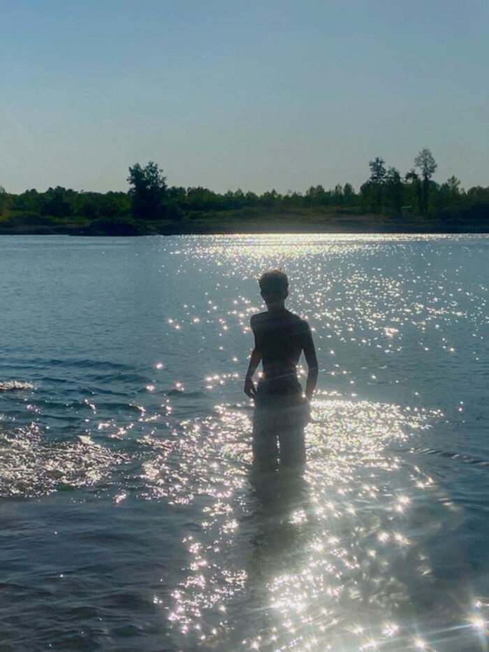 A person standing waist-deep in a lake, silhouetted against a low sun scattering light across the water.
